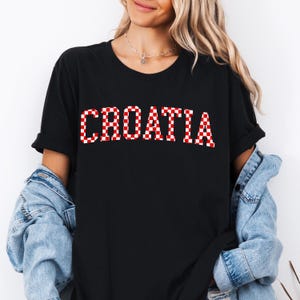 May include: Black t-shirt with the word "CROATIA" in large, red and white checkered letters. The shirt has short sleeves and a crew neck. A denim jacket is worn over the shirt.