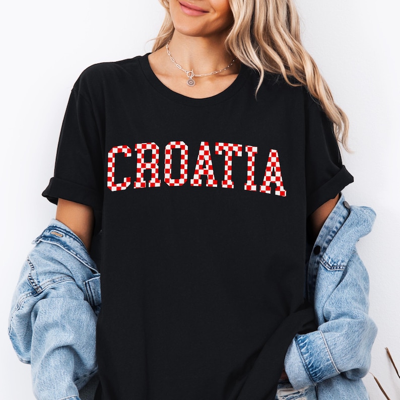 May include: Black t-shirt with the word "CROATIA" in large, red and white checkered letters. The shirt has short sleeves and a crew neck. A denim jacket is worn over the shirt.