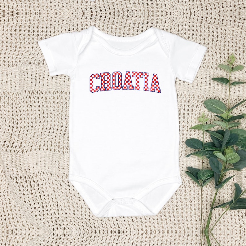 May include: White baby bodysuit with the word "CROATIA" in red and white checkered letters. The bodysuit has short sleeves and a round neckline. The text is in a bold, playful font, and the overall design is simple and clean.