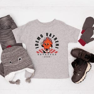 May include: A light gray toddler t-shirt with a cartoon flame character and the text "IDEMO VATRENI HRVATSKA -2026-". The shirt is surrounded by a gray beanie, gray jeans, brown boots, and gray mittens.