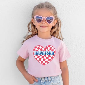 Puede incluir: Una camiseta rosa con un dise&ntilde;o de coraz&oacute;n a cuadros rojos y blancos. El coraz&oacute;n tiene la palabra "HRVATSKA" escrita en letras azules en su interior.