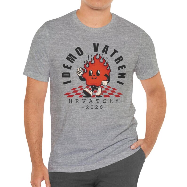 May include: A heather gray t-shirt featuring a cartoon flame character with the text "IDEMO VATRENI" and "HRVATSKA - 2026". The flame is red with a peace sign and is standing on a red and white checkered pattern.