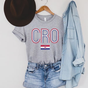 May include: A gray t-shirt with the word "CRO" in blue and red letters with a white outline. The Croatian flag is below the word. The shirt is on a hanger with a denim shirt and a brown hat in the background.