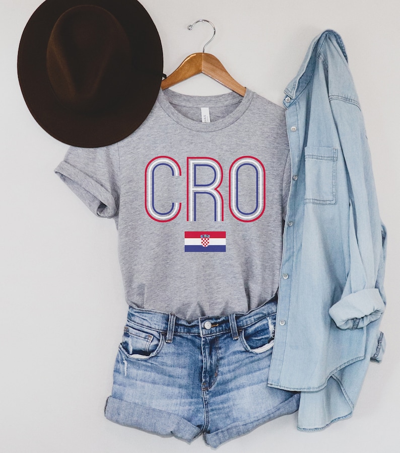 May include: A gray t-shirt with the word "CRO" in blue and red letters with a white outline. The Croatian flag is below the word. The shirt is on a hanger with a denim shirt and a brown hat in the background.