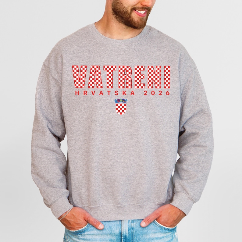 May include: A light grey sweatshirt featuring the word "VATRENI" in red checkered letters. Below the word is "HRVATSKA 2026" in red letters, and a Croatian coat of arms. The sweatshirt has long sleeves and a crew neck.
