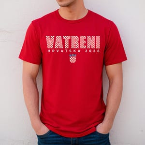 May include: A red t-shirt with the word "VATRENI" in white checkered letters. Below the word is "HRVATSKA 2026" in white letters, and a Croatian coat of arms. The shirt is short-sleeved.