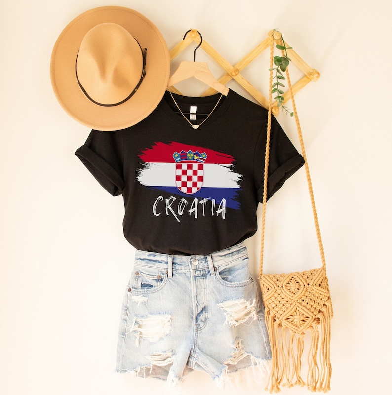 K&ouml;nnte beinhalten: Schwarzes T-Shirt mit einer wei&szlig;en und roten kroatischen Flaggengrafik und dem darunter gedruckten Wort "CROATIA". Das Shirt h&auml;ngt an einem Holzgestell mit einem braunen Hut und einer Strohtasche.