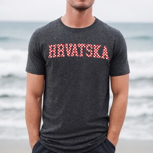 May include: Dark grey t-shirt featuring the word "HRVATSKA" in a red and white chequered pattern. The short-sleeved shirt is made of a soft material. The background shows a blurred view of the sea.