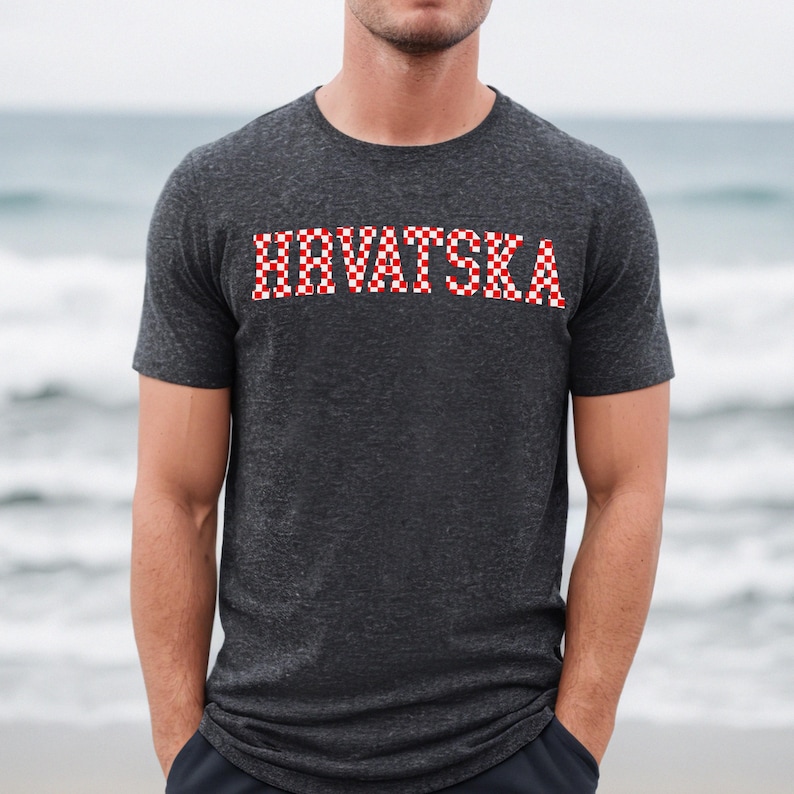 May include: Dark grey t-shirt featuring the word "HRVATSKA" in a red and white chequered pattern. The short-sleeved shirt is made of a soft material. The background shows a blurred view of the sea.
