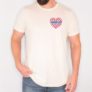 Puede incluir: Una camiseta blanca con un coraz&oacute;n a cuadros rojo y blanco que dice "CROATIA" en letras azules.