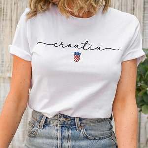 May include: White t-shirt with the word "croatia" written in cursive script and a red and white checkered shield with blue accents.