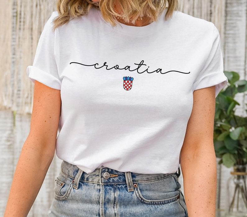 May include: White t-shirt with the word "croatia" written in cursive script and a red and white checkered shield with blue accents.