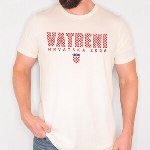 May include: Cream-colored t-shirt featuring the word "VATRENI" in a red checkered pattern, with "HRVATSKA 2026" below. A Croatian coat of arms is centered below the text. The shirt is short-sleeved.