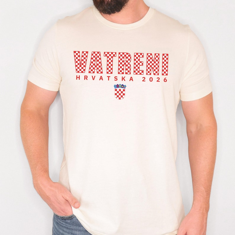 May include: Cream-colored t-shirt featuring the word "VATRENI" in a red checkered pattern, with "HRVATSKA 2026" below. A Croatian coat of arms is centered below the text. The shirt is short-sleeved.
