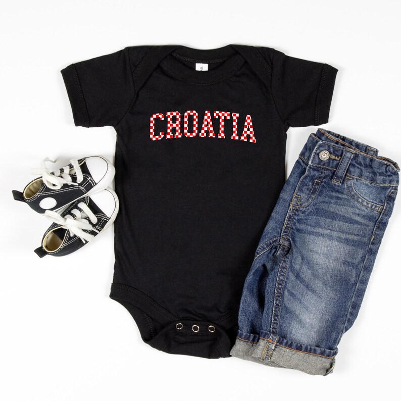 May include: A black baby onesie with the word "CROATIA" in red and white checkered letters. The onesie is paired with a pair of denim jeans and black and white baby sneakers.