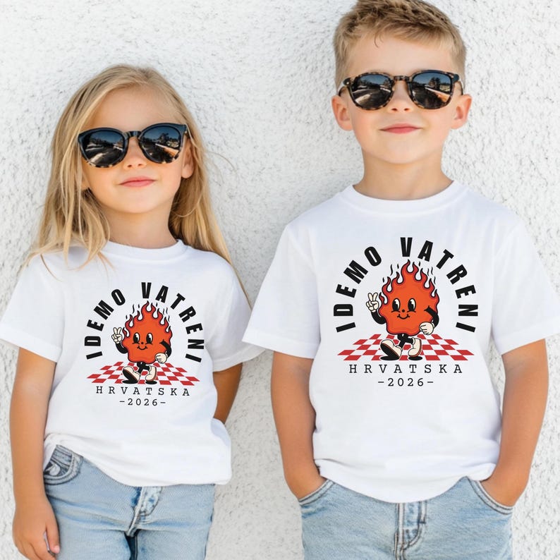 May include: Two children wearing white t-shirts with a cartoon flame design. The design includes the text "IDEMO VATRENI" and "HRVATSKA -2026-" in black. The flame character is red with a peace sign. The shirts have a checkered pattern.