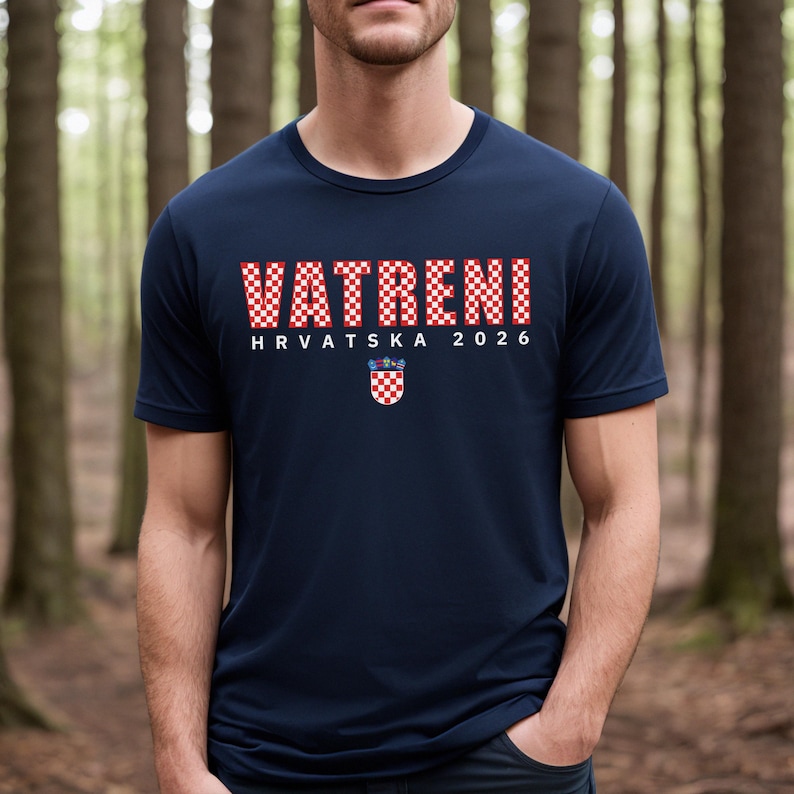 May include: Navy blue t-shirt with the word "VATRENI" in red and white checkered letters. Below the text is "HRVATSKA 2026" and a Croatian coat of arms. The shirt is a crew neck style.