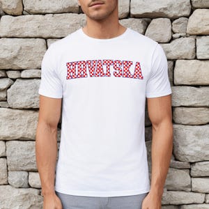 May include: White t-shirt featuring the word "HRVATSKA" in a red and blue chequered design. The shirt has short sleeves and a crew neck. The text is in a bold, blocky font.
