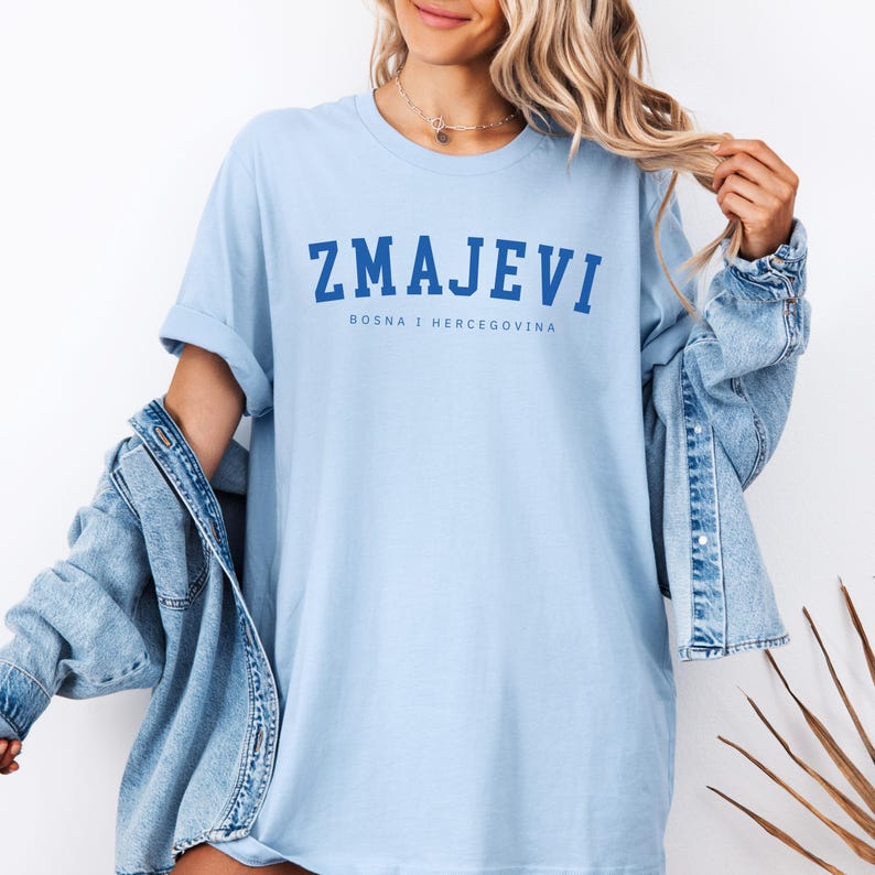 Op de afbeelding: Een lichtblauw t-shirt met de tekst "ZMAJEVI" in blauw, en de tekst "BOSNA I HERCEGOVINA" in kleinere blauwe letters eronder.