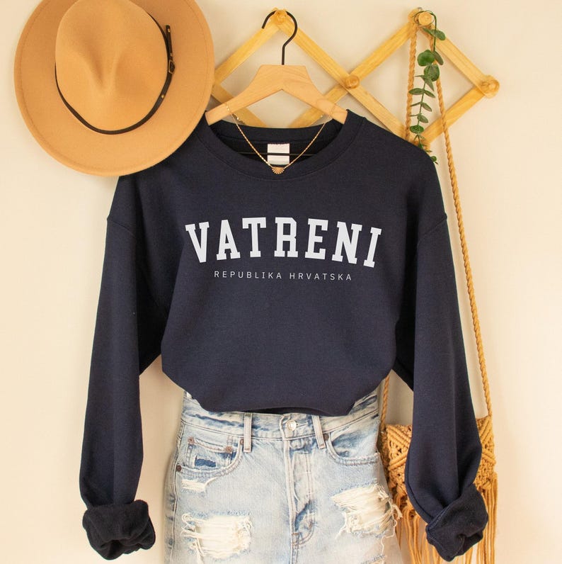 Pu&ograve; includere: Una felpa blu navy con testo bianco che recita "VATRENI REPUBLIKA HRVATSKA". La felpa &egrave; appesa a un supporto in legno con un cappello marrone e una borsa di paglia.