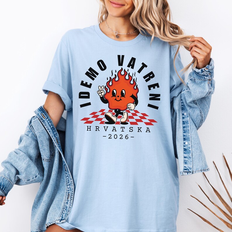 Puede incluir: Camiseta azul claro con un personaje de llama de dibujos animados y el texto "IDEMO VATRENI" y "HRVATSKA - 2026-". El dise&ntilde;o incluye un patr&oacute;n de cuadros rojos y blancos. La camiseta tiene un corte holgado.