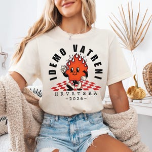 Puede incluir: Camiseta color crema con un personaje de llama de dibujos animados haciendo el signo de la paz. El texto "IDEMO VATRENI" est&aacute; encima de la llama, con "HRVATSKA - 2026-" debajo. La llama est&aacute; sobre un patr&oacute;n de cuadros rojos y blancos.