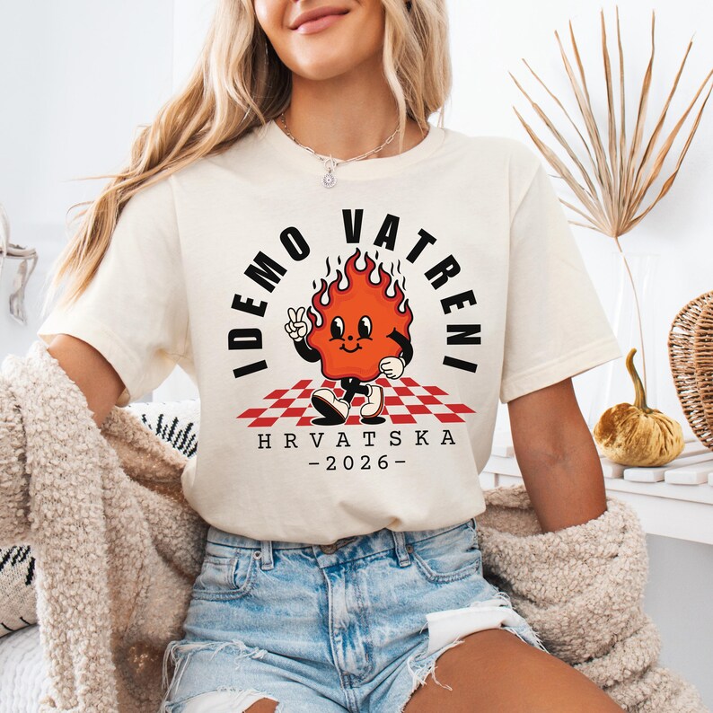 Puede incluir: Camiseta color crema con un personaje de llama de dibujos animados haciendo el signo de la paz. El texto "IDEMO VATRENI" est&aacute; encima de la llama, con "HRVATSKA - 2026-" debajo. La llama est&aacute; sobre un patr&oacute;n de cuadros rojos y blancos.