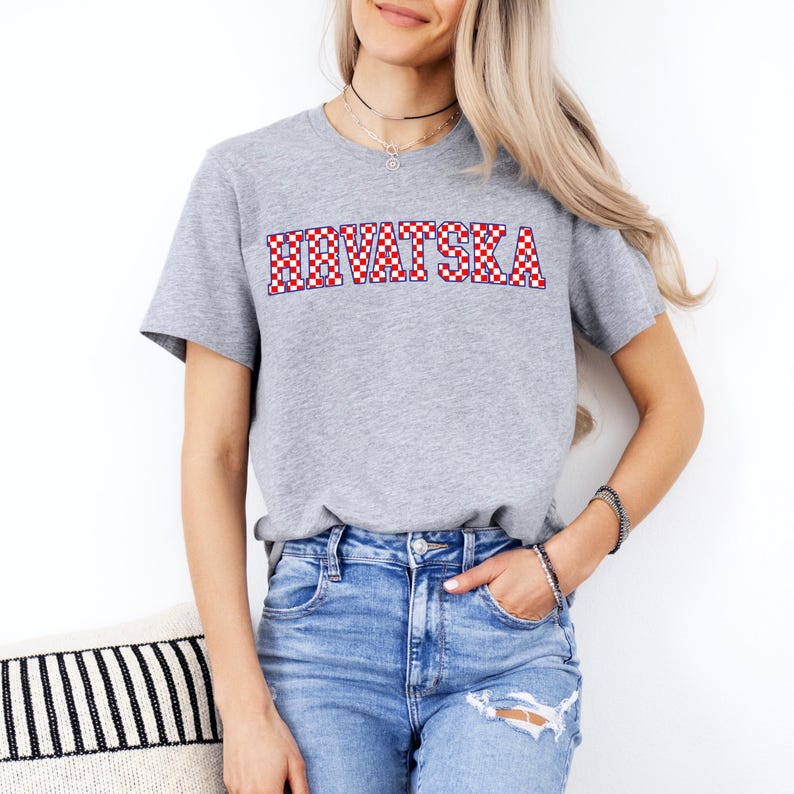 Pode incluir: Camiseta cinza com a palavra "HRVATSKA" em letras xadrez vermelhas e brancas. A camiseta tem mangas curtas e gola redonda. A modelo est&aacute; usando jeans azuis.