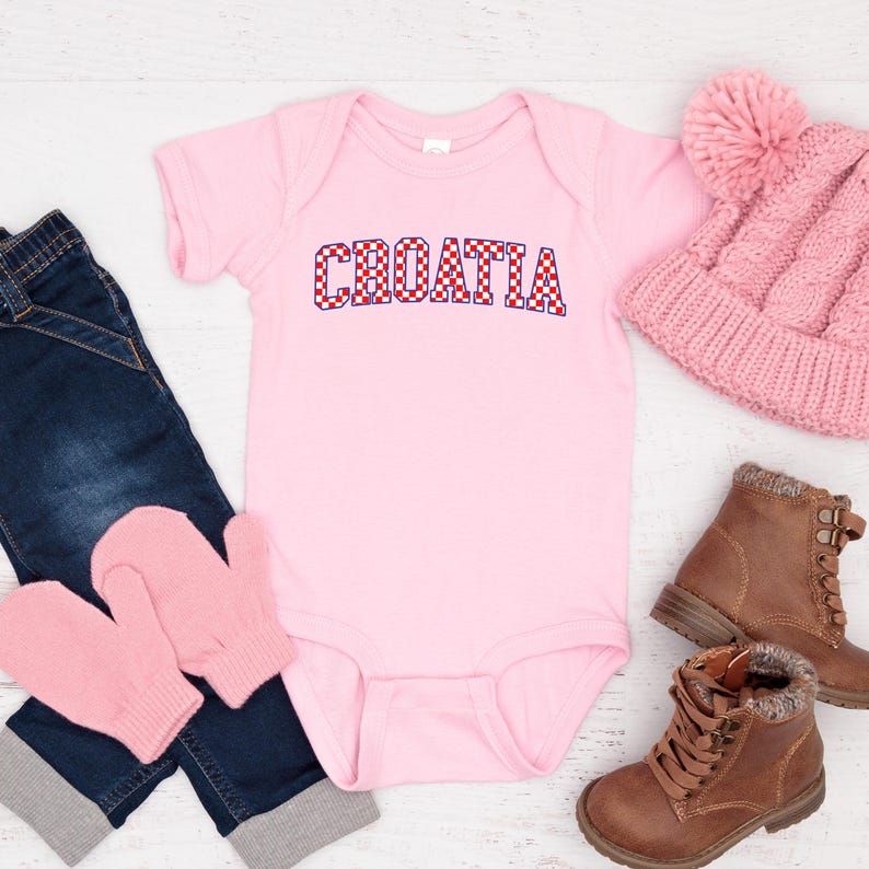 May include: A light pink baby onesie with the word "CROATIA" in a red and white checkered pattern. The onesie is paired with dark blue jeans, pink mittens, a pink knit hat, and brown leather boots.