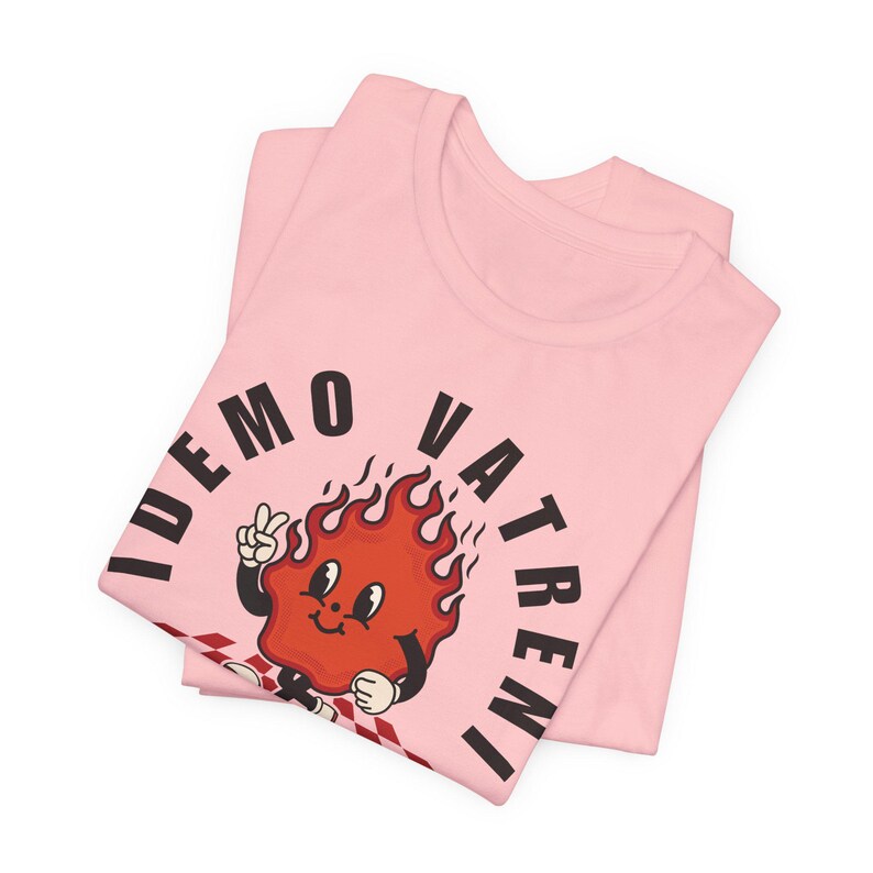 Puede incluir: Dos camisetas de color rosa claro dobladas y apiladas. La camiseta superior presenta un personaje de llama de dibujos animados con el texto "IDEMO VATRENI" en negro. El personaje de la llama es rojo con un signo de paz blanco y un patr&oacute;n de cuadros.