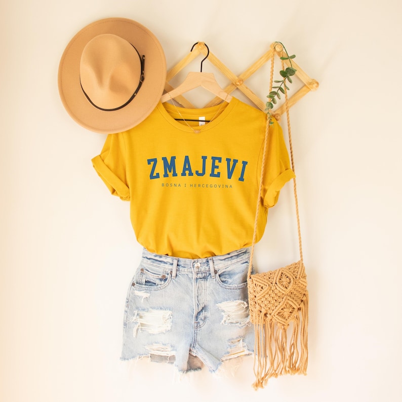Op de afbeelding: Een geel T-shirt met de tekst "ZMAJEVI" in blauwe letters en de woorden "BOSNA I HERCEGOVINA" in kleinere blauwe letters eronder. Het shirt hangt aan een houten rek met een strohoed en een macram&eacute; tas.
