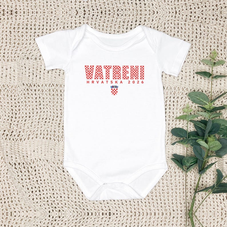 K&ouml;nnte beinhalten: Wei&szlig;er Baby-Body mit kurzen &Auml;rmeln. Auf der Vorderseite steht das Wort "VATRENI" in rot karierten Buchstaben, darunter "HRVATSKA 2026". Ein kleines kroatisches Wappen ist ebenfalls auf der Vorderseite aufgedruckt.