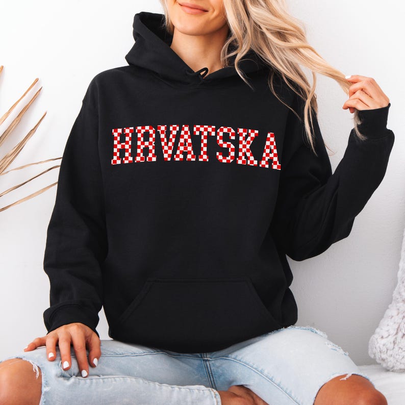 May include: Black hooded sweatshirt with the word "HRVATSKA" in red and white checkered letters across the chest. The sweatshirt has a front pocket and a drawstring hood. The sleeves are long and the material appears to be a soft fabric.