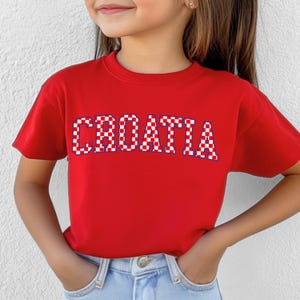 May include: A red t-shirt with the word "CROATIA" in white letters with a red and white checkered pattern. The shirt is being worn by a person.
