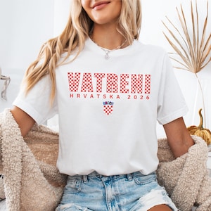 May include: White t-shirt with the word "VATRENI" in red checkered letters. Below the word are the words "HRVATSKA 2026" and a Croatian coat of arms. The shirt is made of a soft material.