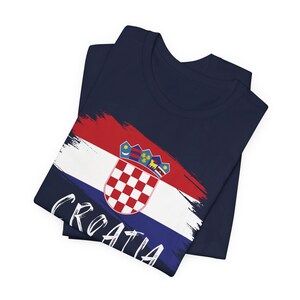 K&ouml;nnte beinhalten: Ein marineblaues T-Shirt mit einem kroatischen Flaggen-Design in Wei&szlig; und Rot. Die Flagge zeigt einen wei&szlig;en Schild mit einem Schachbrettmuster und einer Krone. Das Wort "CROATIA" ist in Wei&szlig; am unteren Rand des Shirts geschrieben.