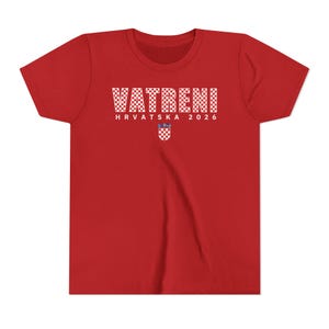 May include: A red t-shirt with the word "VATRENI" in white checkered letters. Below the word is "HRVATSKA 2026" in white letters, and a Croatian coat of arms. The shirt is made of a soft material.