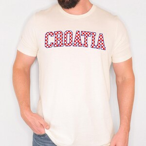 May include: Cream-colored t-shirt with the word "CROATIA" in a red and white checkered pattern. The text is outlined in blue. The shirt has short sleeves and a crew neck.