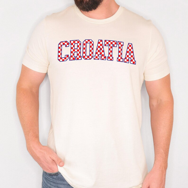 May include: Cream-colored t-shirt with the word "CROATIA" in a red and white checkered pattern. The text is outlined in blue. The shirt has short sleeves and a crew neck.