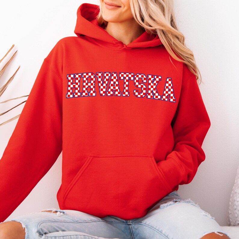 May include: A red hooded sweatshirt with the word "HRVATSKA" in a checkered pattern across the chest. The sweatshirt has a front pocket and a drawstring hood. The person is wearing blue jeans.