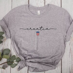 May include: Gray t-shirt with the word "croatia" written in cursive script and a small Croatian coat of arms below it.