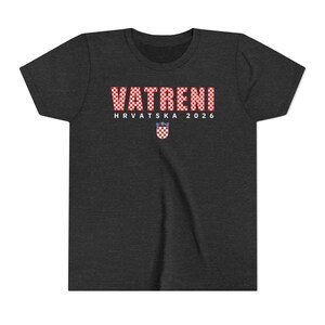 May include: Dark gray t-shirt with the word "VATRENI" in a red and white checkered pattern. Below the word is "HRVATSKA 2026" and a Croatian coat of arms. The shirt is made of a soft material.