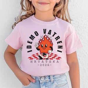May include: A light pink t-shirt featuring a cartoon flame character with the text "IDEMO VATRENI" and "HRVATSKA - 2026-". The flame is on a red and white checkered pattern.
