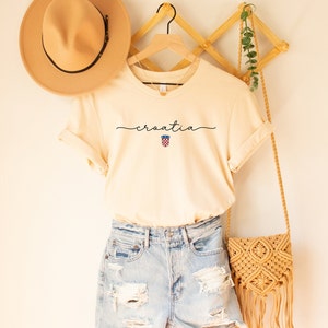 May include: A cream colored t-shirt with the word "croatia" written in cursive with a small Croatian flag symbol underneath. The shirt is hanging on a wooden hanger with a straw hat and a woven bag.