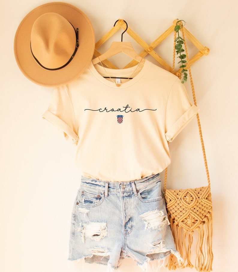 May include: A cream colored t-shirt with the word "croatia" written in cursive with a small Croatian flag symbol underneath. The shirt is hanging on a wooden hanger with a straw hat and a woven bag.