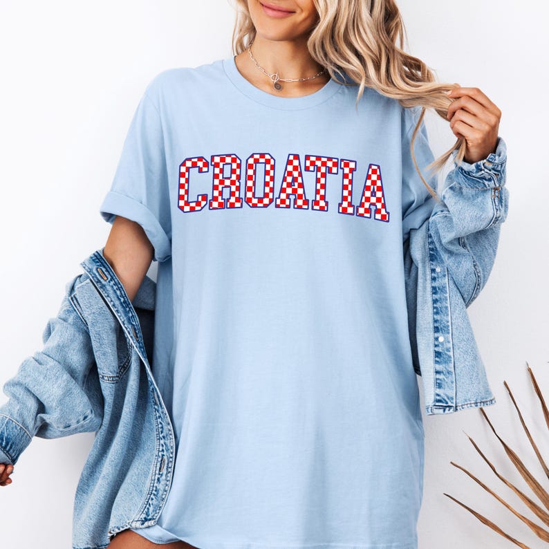 May include: Light blue t-shirt with the word "CROATIA" in red and white checkered letters. The shirt has short sleeves and a relaxed fit. A denim jacket is worn over the shirt.