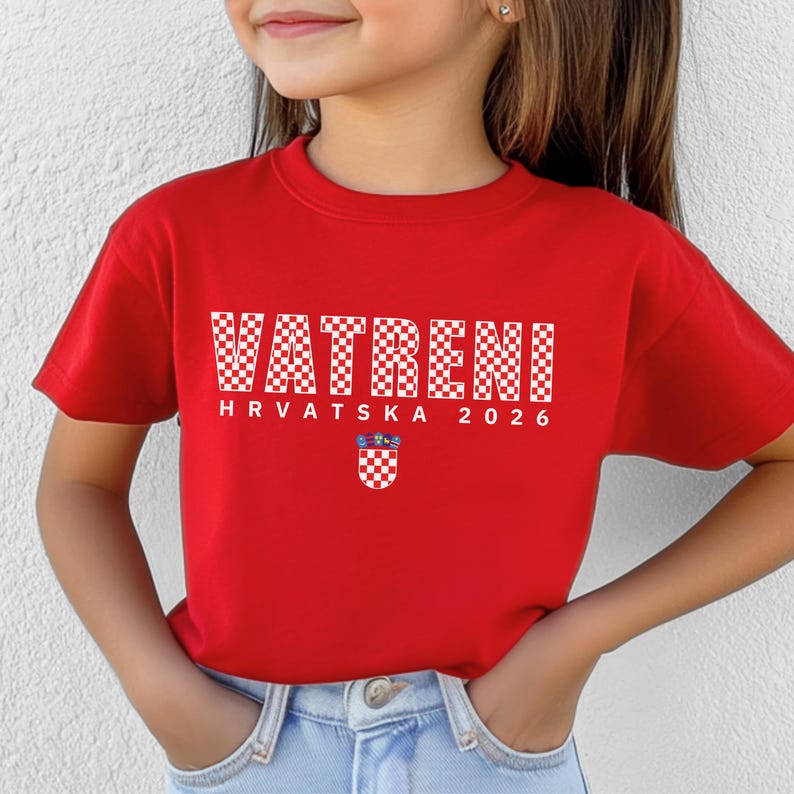 May include: A red t-shirt with the word "VATRENI" in a white checkered pattern. Below the word is "HRVATSKA 2026" and a Croatian coat of arms. The shirt is short-sleeved.