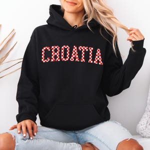 May include: Black hooded sweatshirt with the word "CROATIA" in red and white checkered letters across the chest. The sweatshirt has a front pocket and a drawstring hood. The model is wearing blue jeans.