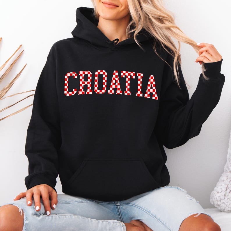 May include: Black hooded sweatshirt with the word "CROATIA" in red and white checkered letters across the chest. The sweatshirt has a front pocket and a drawstring hood. The model is wearing blue jeans.