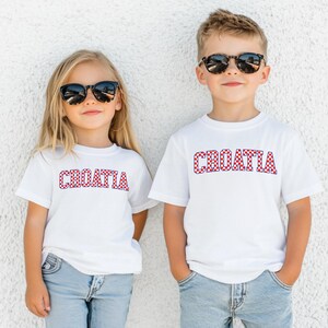 May include: Two children wearing white t-shirts with the word "CROATIA" in red and white checkered letters. Both children are wearing sunglasses and blue jeans. The background is a plain white wall.